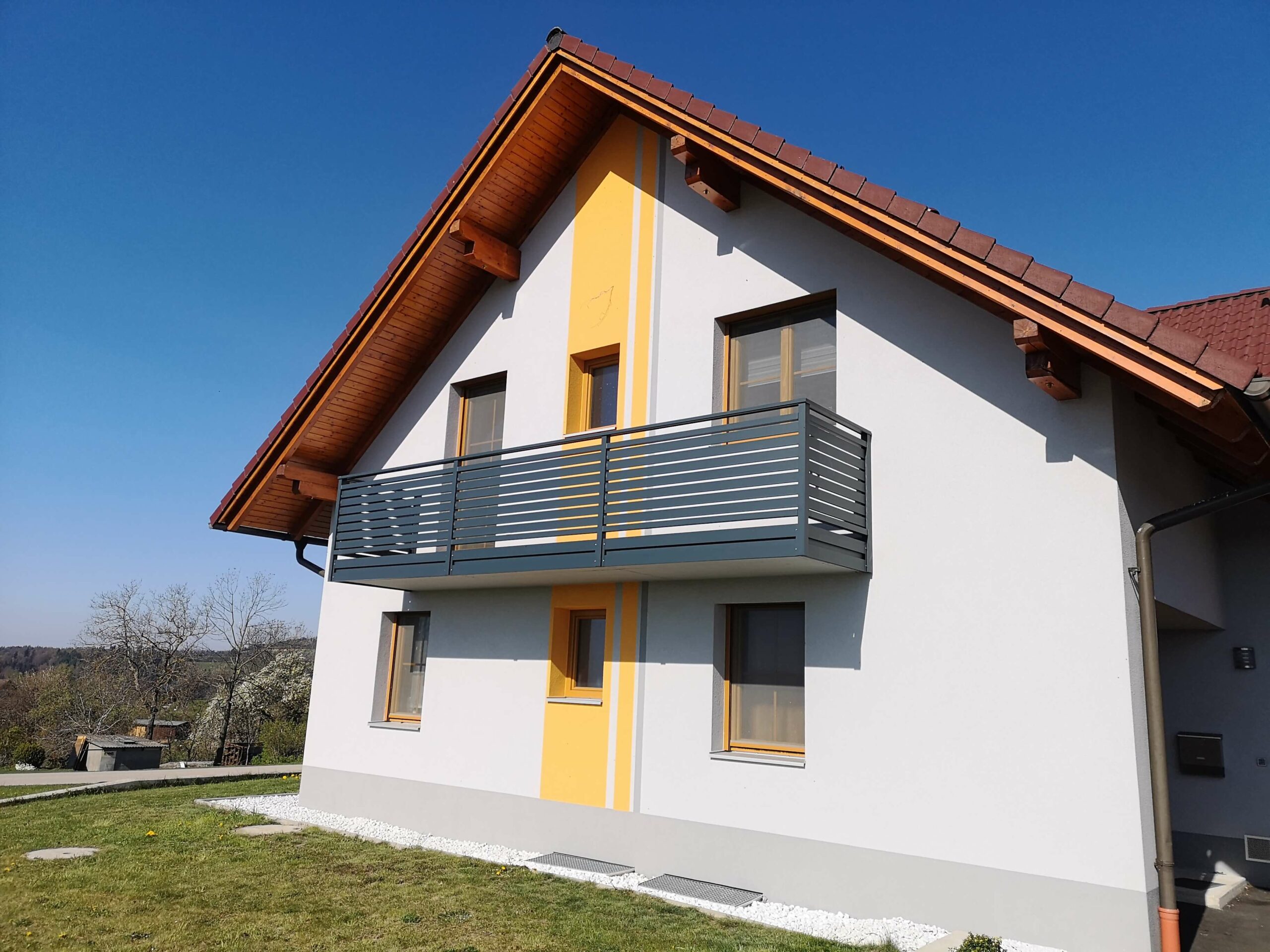 Grauer Alubalkon an einem Haus mit weißer Fassade mir orangenen Elementen